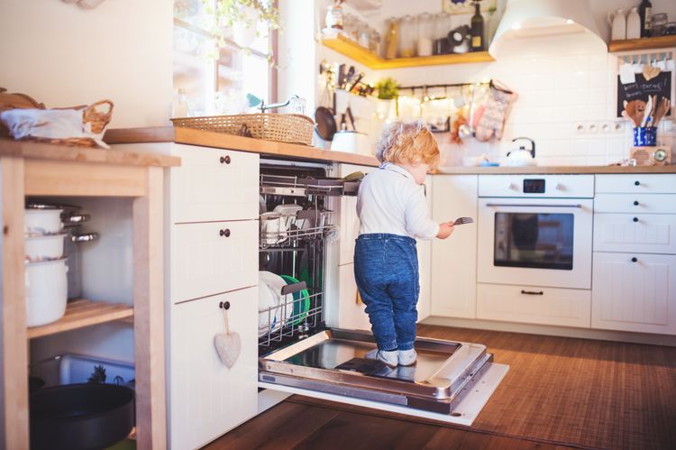 Kleinkind steht auf offener Geschirrspülertüre in weißer Einbauküche