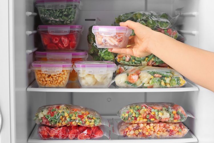 Frauenhand entnimmt Kunststoffbox mit Gefriergut aus Tiefkühlschrank, dahinter weitere Boxen mit buntem gefrorenem Gemüse