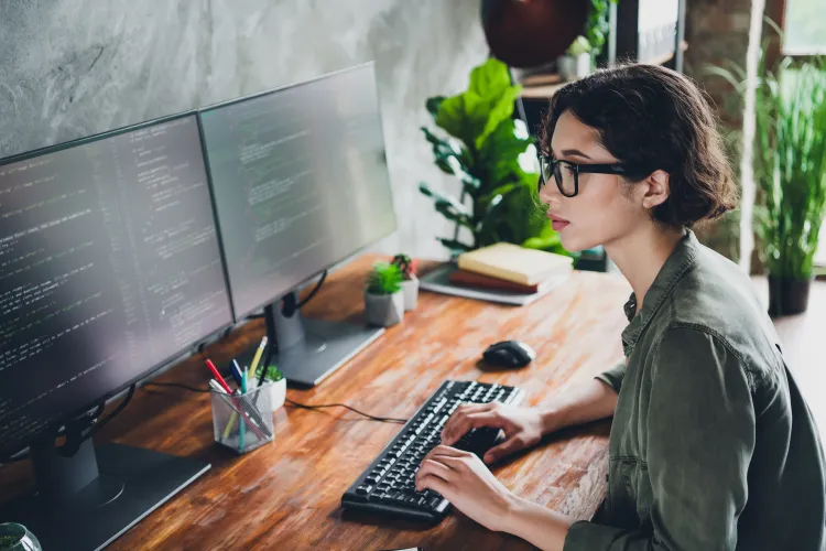 Junge Frau schaut auf zwei Computermonitore am Schreibtisch.