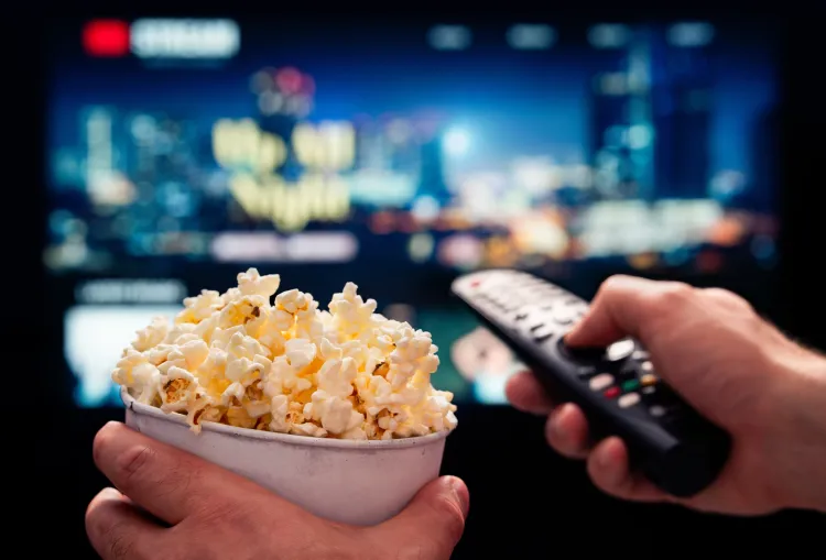 Eine Person bedient einen Fernseher mittels einer Fernbedienung mit einem Eimer Popcorn daneben.
