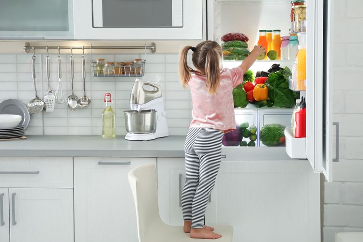 Kind steht auf Stuhl vor offenem Kühlschrank und greift nach Apfel