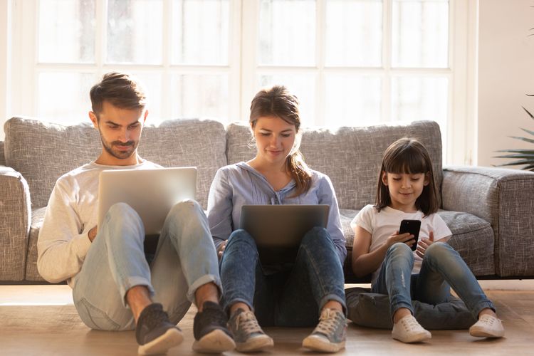 Vater, Mutter und Tochter sitzen vor ihrer Wohnzimmercouch am Boden und benutzen LAptops und Smartphone