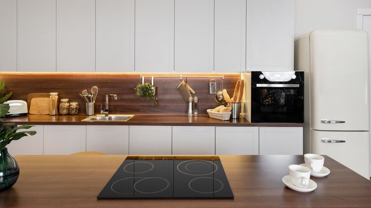 Panoramasicht auf moderne Küche mit leerem Keramikkochfeld auf einer Holztheke vor weißen Küchenschränken, Waschbecken, Backofen und Kühlschrank