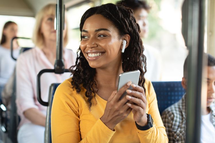 Junge lächelnde Frau beim Telefonieren mit Ohrhörern im Bus, in der Hand ihr Smartphone