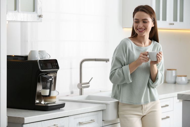 Junge Frau in weißer Einbauküche trinkt Kaffee aus Vollautomaten mit zufriedenem Blick aufs Gerät