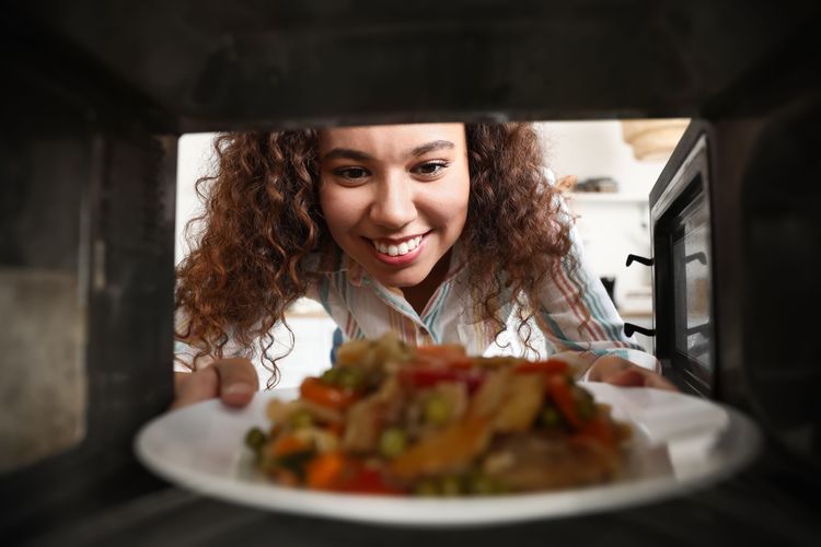 lachende junge Frau mit brünettem Lockenkopf blickt in Mikrowelle und holt Teller mit Essen raus