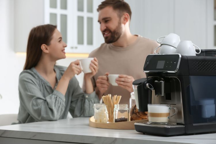 Paar in Küche hält Kaffeetassen in den Händen und lächelt sich an, daneben ein Kaffeevollautomat