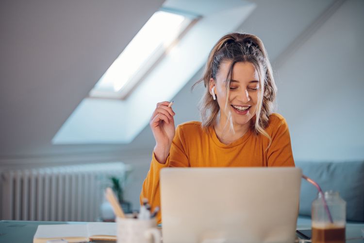 Junge blonde Frau spricht lächelend mit In-Ear-Headset an ihrem Schreibtisch vor dem Laptop