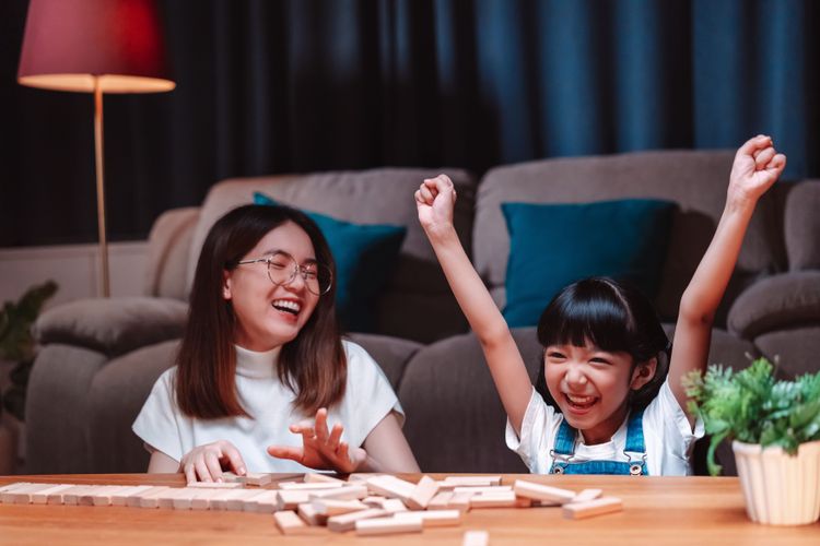 Kleines asiatisches Mädchen hat Brettspiel gegen Mutter gewonnen und reißt freudig lachend die Arme nach oben