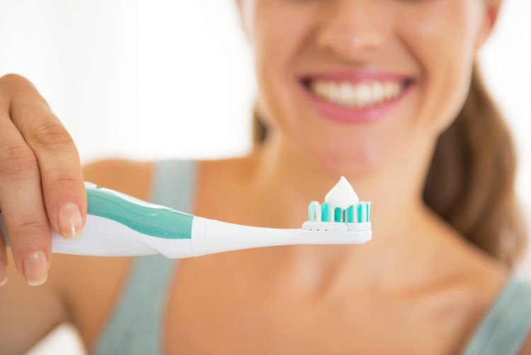 Weibliche Hand hält Elektrozahnbürste mit Zahnpasta, lächelnde Frau im Hintergrund
