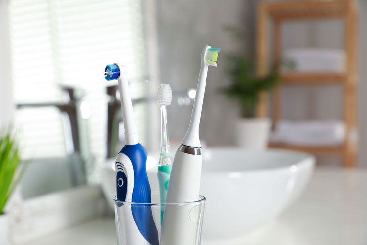 Schallzahnbürste, rotierende Zahnbürste und Kinder-Elektrozahnbürste im Zahnputzbecher, im Hintergrund ein Badezimmer