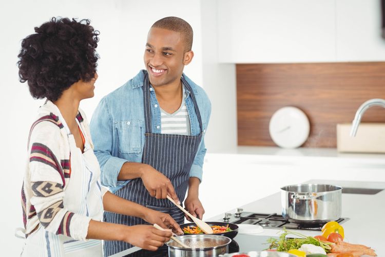 Mann lächelt seine Frau an während beide in moderner Küche Gemüsegericht auf Gas und Glaskeramik zubereiten