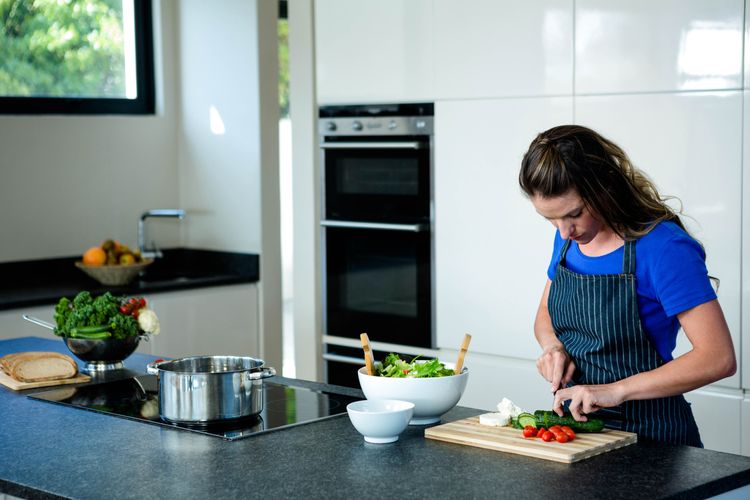 Frau schneidet auf einer Kochinsel mit Kochfeld Gemüse, im Hintergrund eine weiße Einbauküche mit Backrohr