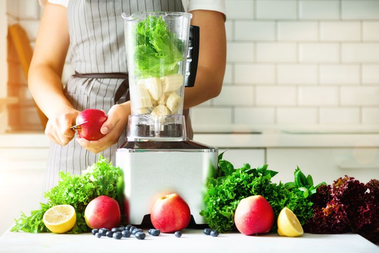 Food Processor gefüllt mit grünem Gemüse, dahinter schneidet eine Frau einen Apfel, rundherum liegt weiteres Obst und Gemüse